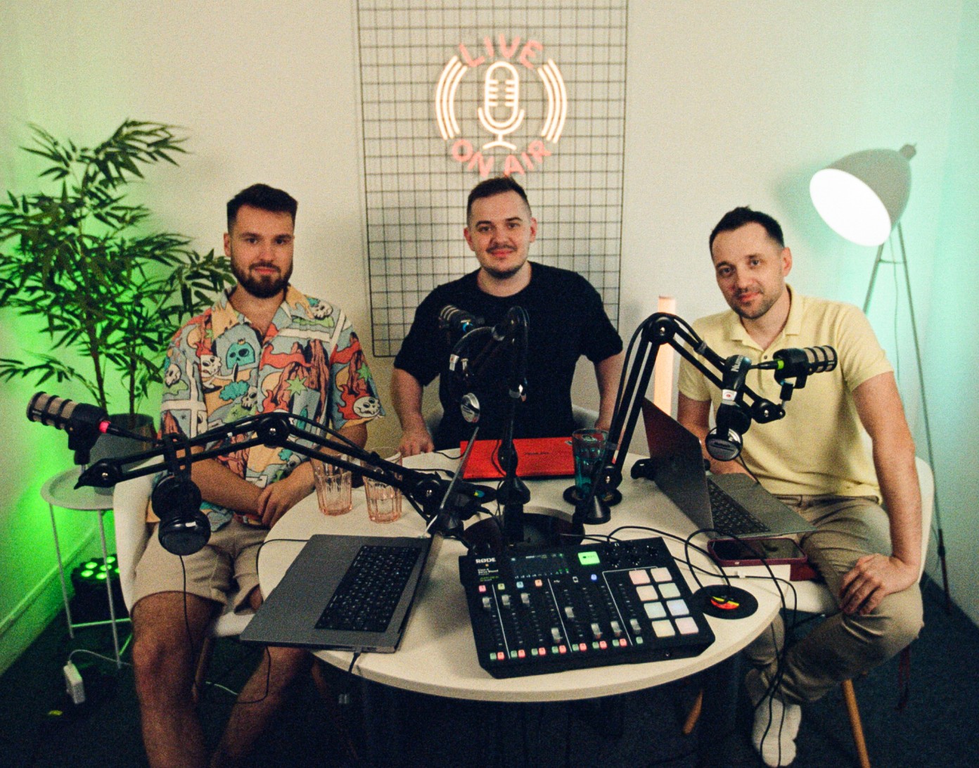 Three men at a podcast table with microphones, laptops, and a mixing board. A neon sign above says "Live On Air".
