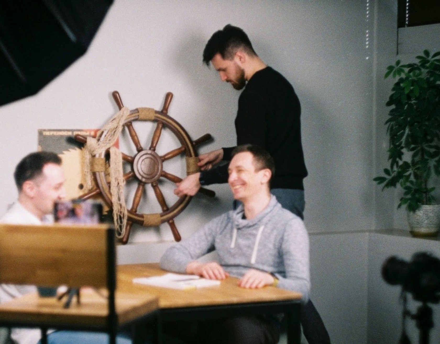 Three men with a ship steering wheel prop, two seated, one standing, indoors. One man smiles while another touches the wheel.